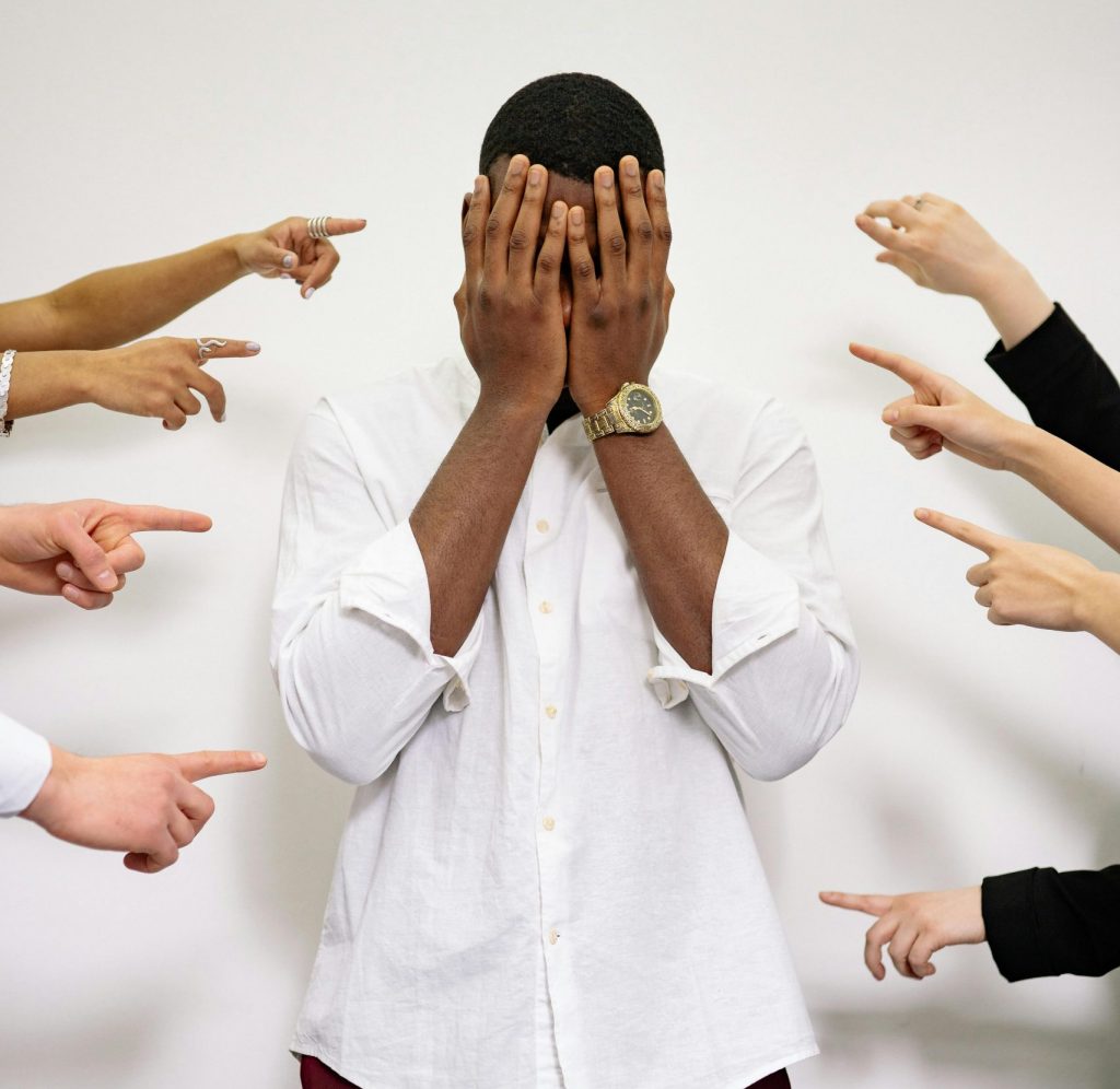 A man in distress covers his face surrounded by multiple pointing fingers.